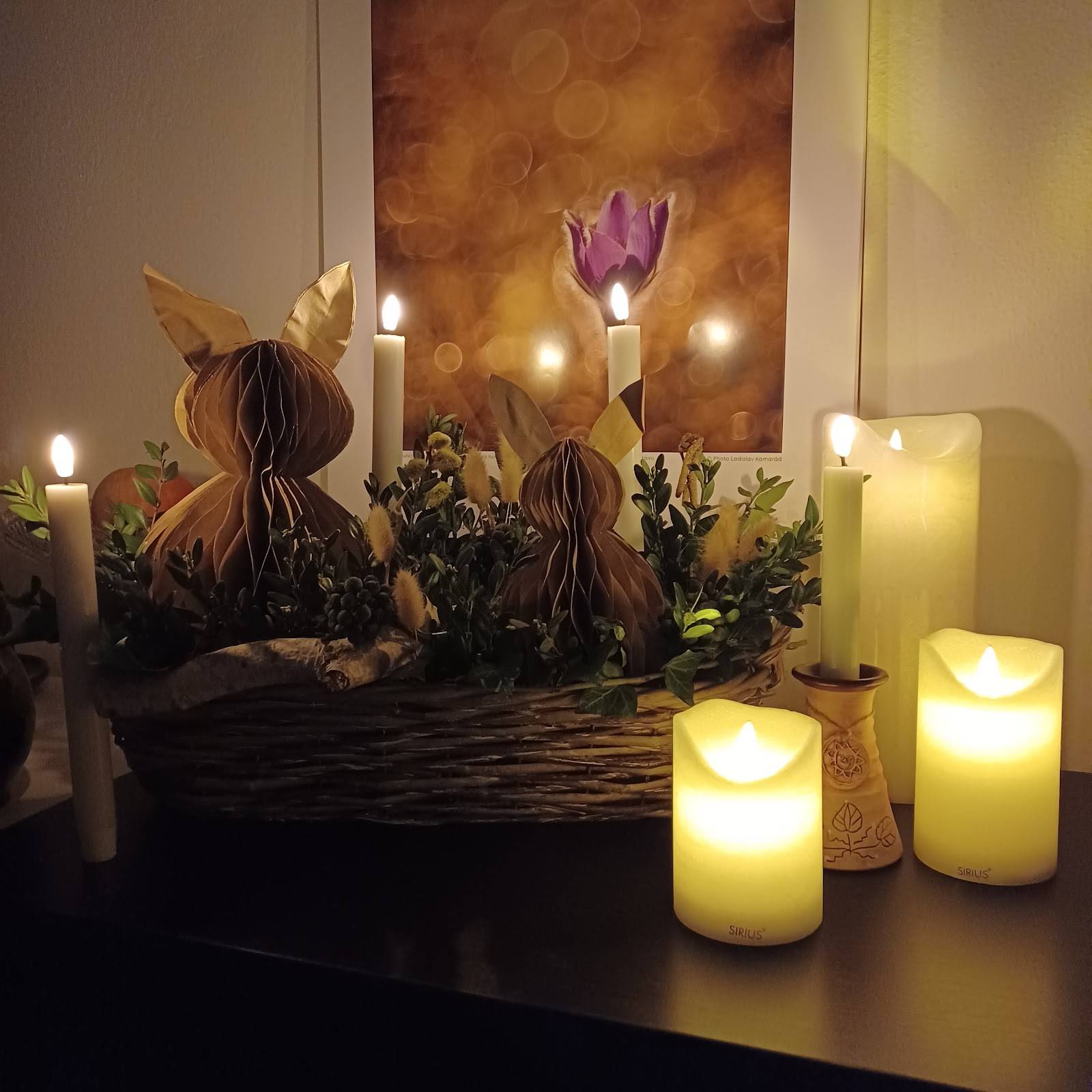 Detailní pohled na velikonoční aranžmá se zajíčky a hřejivým světlem svíček v proutěném košíku.
